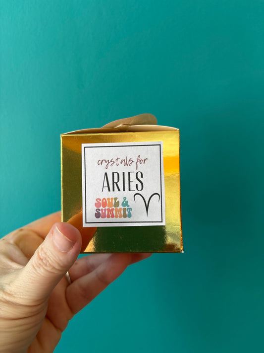 Hand holding gold Aries Zodiac Gift Box with crystals for Aries label against teal background