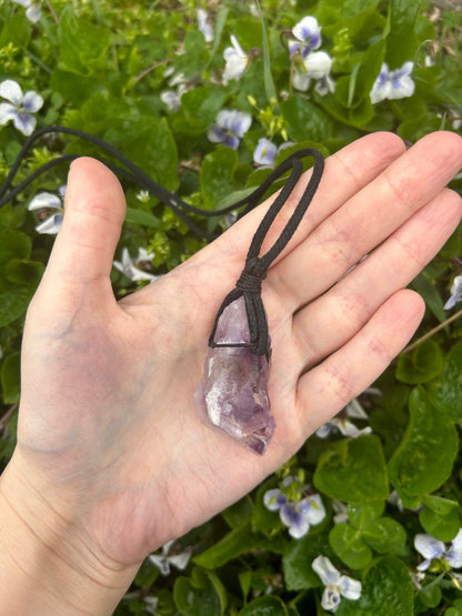 Hand holding a crystal cord necklace with a purple crystal pendant against a background of green leaves and small purple flowers