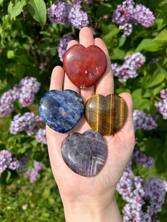 Hand holding four flat crystal hearts in red, blue, golden brown, and purple colors with green leaves and purple flowers in the background