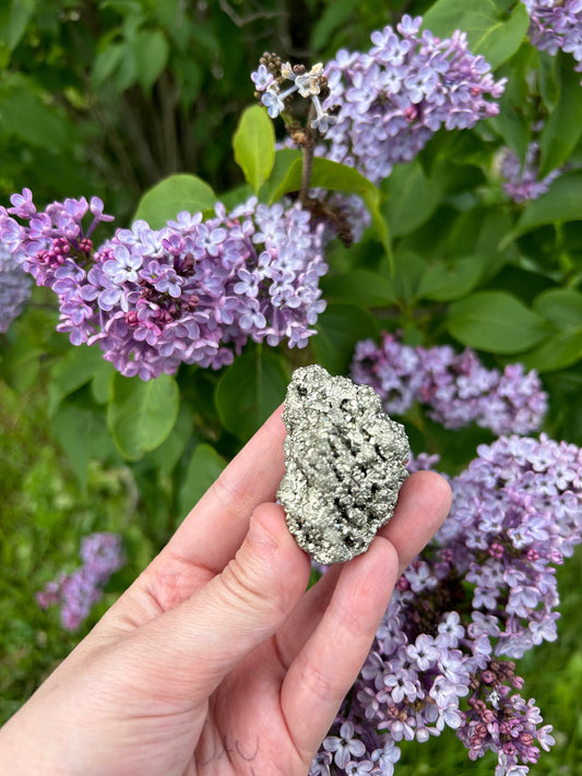 Hand holding a raw pyrite chunk with metallic sheen against a backdrop of blooming purple lilac flowers and green leaves