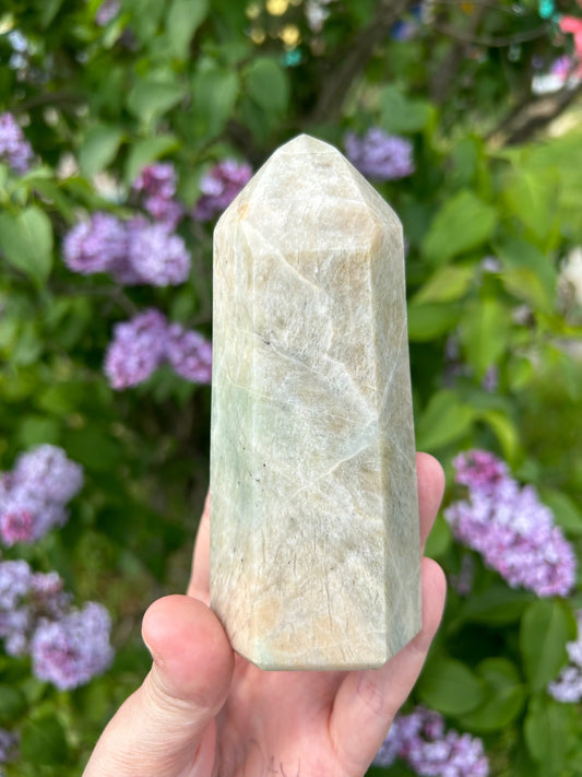 Hand holding a polished Garnierite tower crystal with green and white hues against a background of green leaves and purple flowers
