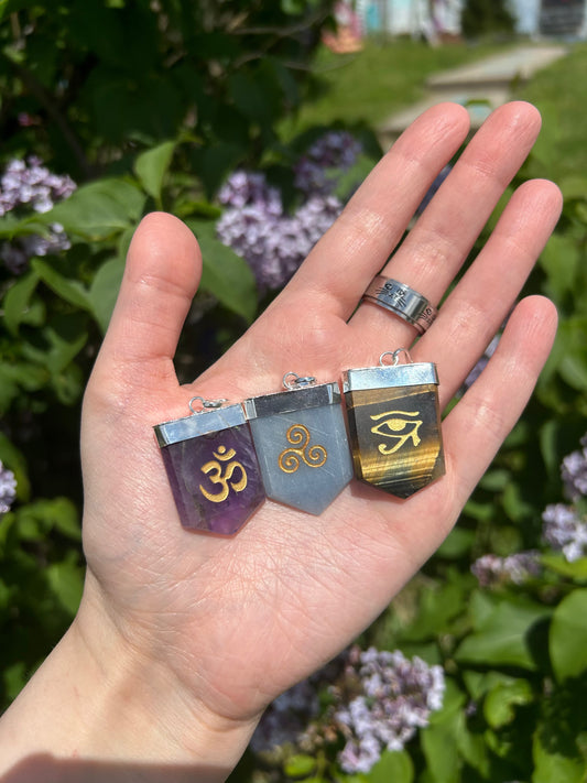 Hand holding three crystal symbol pendants with spiritual engravings outdoors on green foliage background