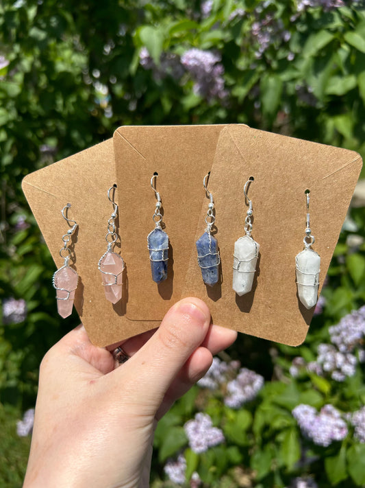 Handcrafted wire wrap crystal earrings in pink, blue, and white displayed on brown cards outdoors with greenery background