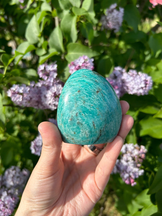 Hand holding a smooth amazonite freeform stone with lilac flowers and green leaves in the background