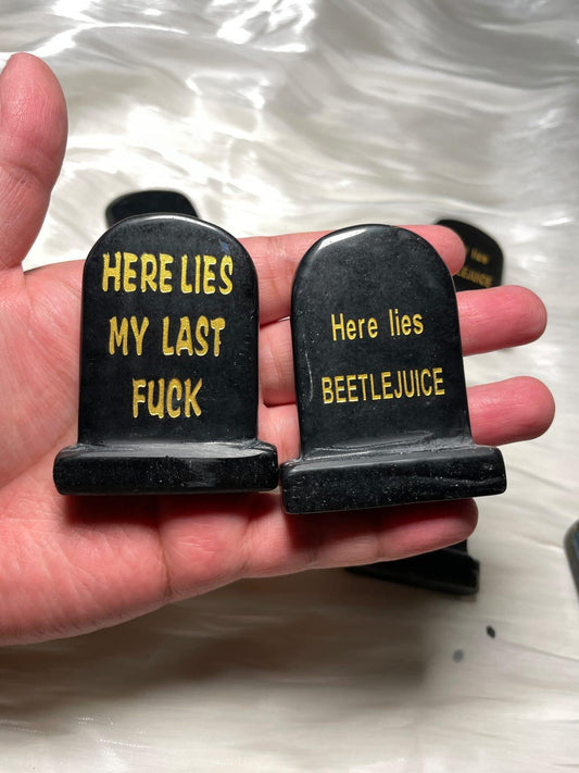 Small black tombstone carvings with gold text, one reads "HERE LIES MY LAST FUCK" and the other "Here lies BEETLEJUICE," held in a hand on a silky white background