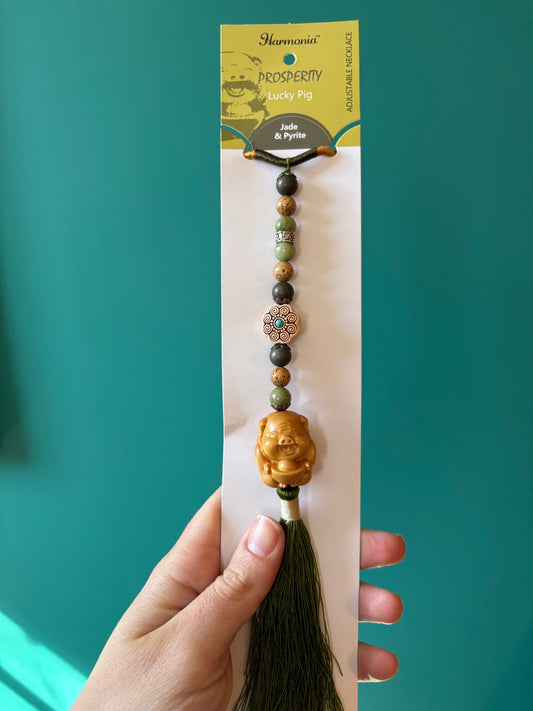 Harmonia necklace with jade, pyrite beads, lucky pig charm, and green fringe tassel hanging on packaging