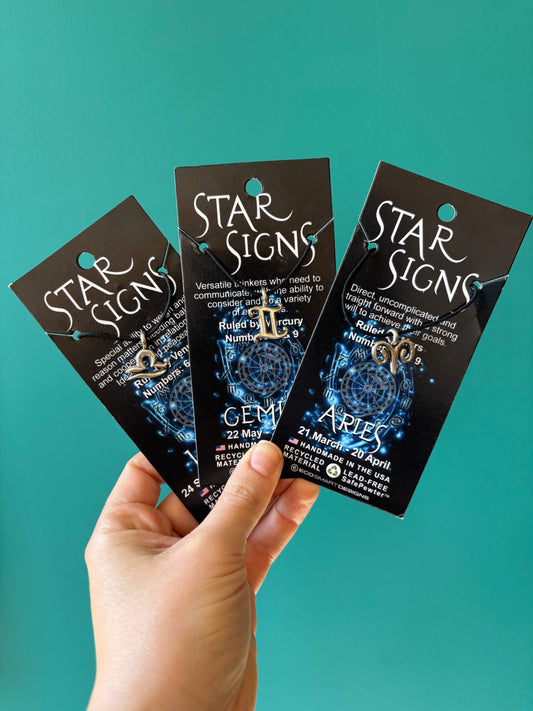 Hand holding three Star Signs zodiac necklaces Aries, Gemini, and Aquarius on black cards against teal background, Zodiac Necklace.