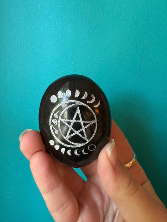 Black tourmaline palm stone with pentacle and moon phase carvings held in hand against turquoise background