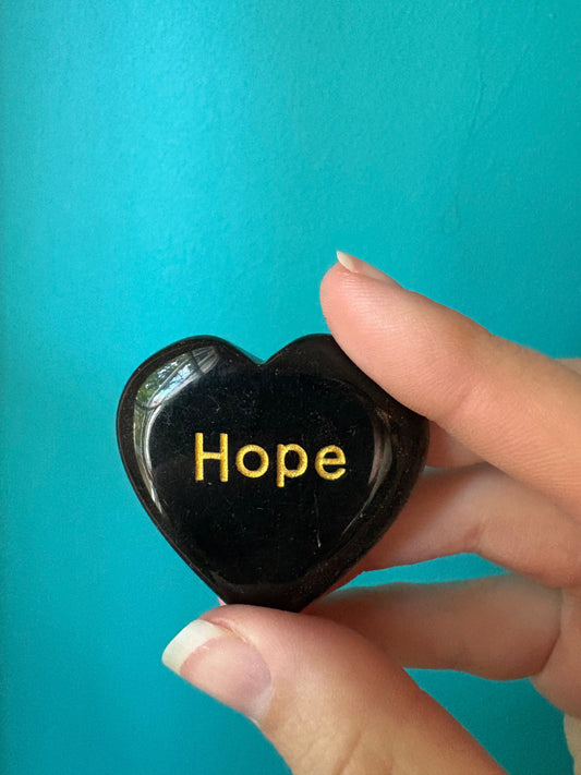Hand holding a smooth black heart-shaped affirmation heart stone engraved with the word Hope against a turquoise background