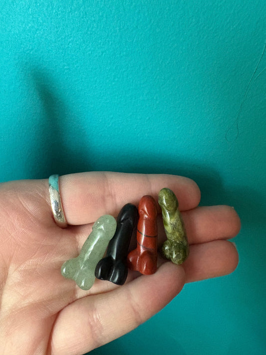 Hand holding four small Teenie Weenie hand-crafted penis figurines in different colors against teal background