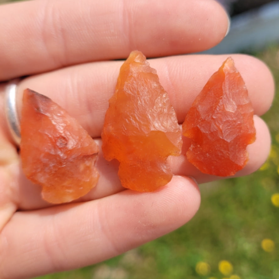 Hand holding three reddish-orange carnelian arrow heads with natural rough texture outdoors