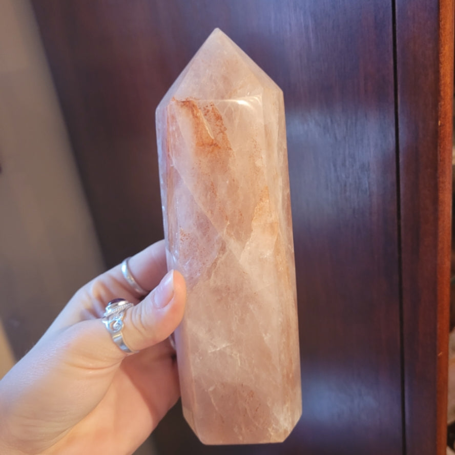 Hand holding a polished hematite quartz tower with natural red and orange inclusions against a wooden background