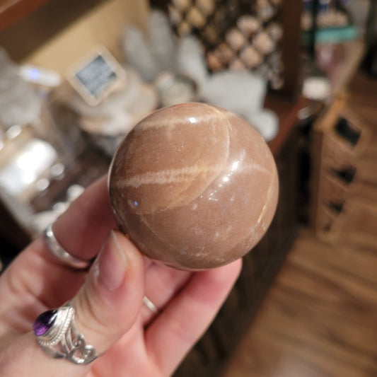 Hand holding a polished peach moonstone sphere with soft peach and cream tones in a blurred indoor setting