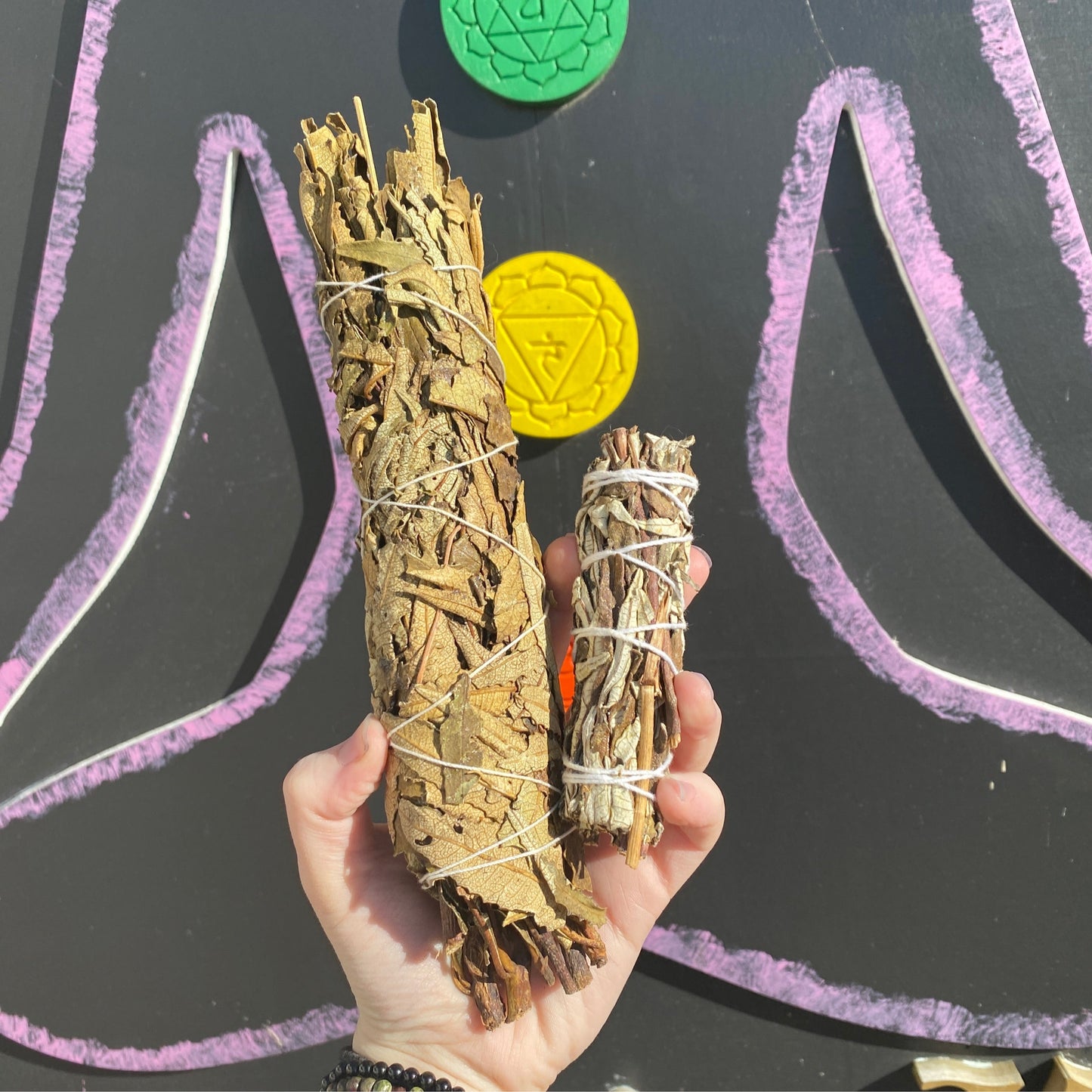 Hand holding two Yerba Santa smudge bundles wrapped with white string against a colorful spiritual background