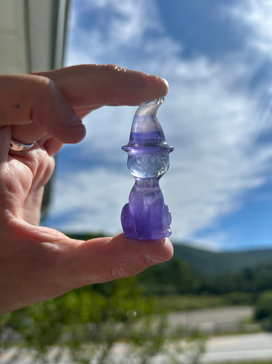 Handheld fluorite cat wizard carving with intricate details and purple hues against a bright sky background