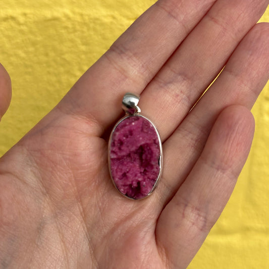 Close-up of a hand holding a pink and purple Cobaltoan Calcite pendant with silver setting against a yellow background