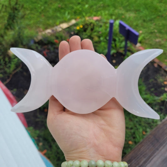 Hand holding a pink calcite triple moon bowl, featuring a smooth central circle with crescent moons on each side, outdoors with greenery in the background