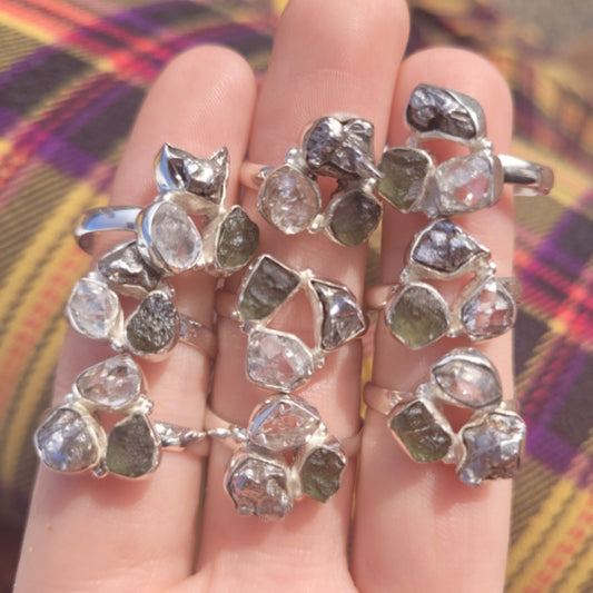 Ring featuring triple stones of moldavite, pakimer diamond, and Campo Del Cielo meteorite on silver bands displayed on fingers.