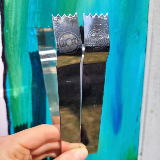 Metal charcoal tongs with serrated edges and engraved symbols on a blue background