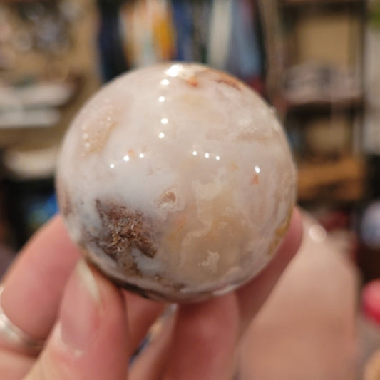 Hand holding a polished flower agate sphere with creamy white and brown floral patterns in a blurred indoor setting