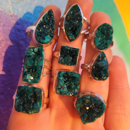 Hand displaying multiple Ring Dioptase Druzy rings with vibrant green-blue crystalline stones in various shapes under sunlight