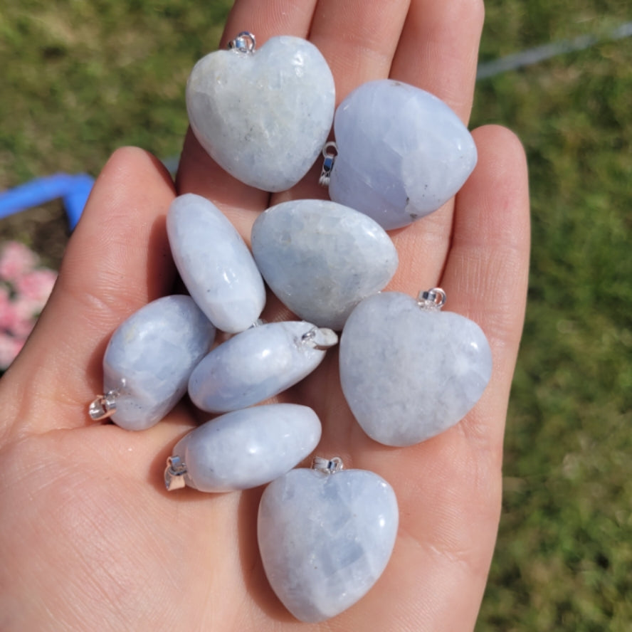 Hand holding multiple heart-shaped blue calcite pendants with silver bails on a blurred outdoor background