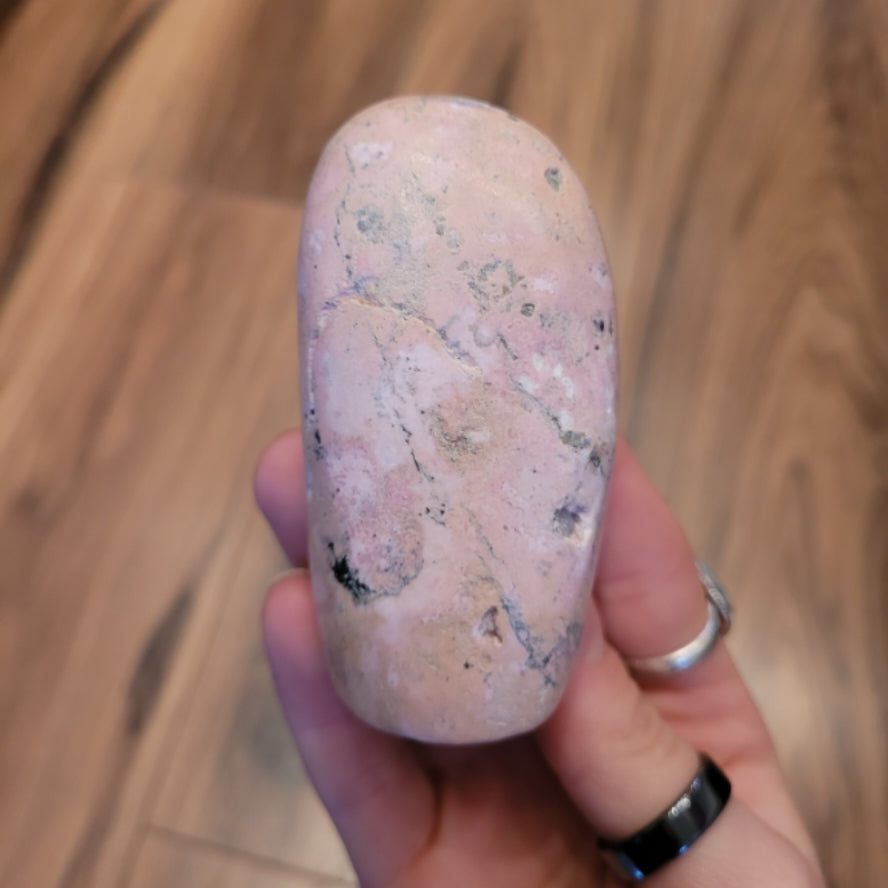 Hand holding a smooth, pink Rhodochrosite freeform stone with natural patterns on a wooden background