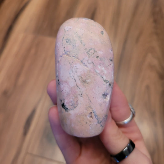 Hand holding a smooth, pink Rhodochrosite freeform stone with natural patterns on a wooden background