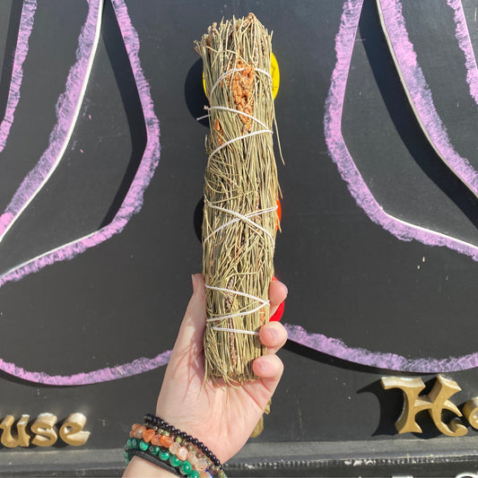 Hand holding a pine smudge bundle wrapped with white string against a dark background with purple and gold decorative elements