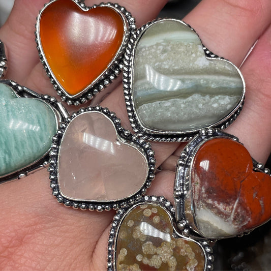 Hand holding silver plated heart shaped rings with various colored stones including orange, green, and beige tones heart shaped ring collection