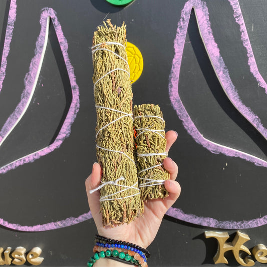Hand holding two juniper smudge bundles wrapped in white string against a blackboard with purple chalk drawings