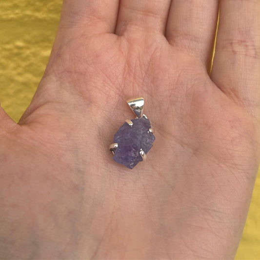 Hand holding a Tanzanite rough pendant with silver setting against a yellow background
