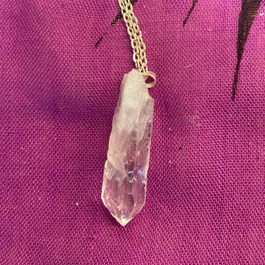 Raw clear quartz pendant on gold chain against purple fabric background