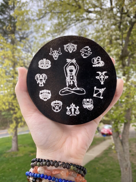 Hand holding black selenite plate engraved with 12 zodiac symbols and meditative figure outdoors with trees in background