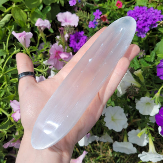 Hand holding a long oval Selenite oval bowl with a smooth translucent white crystal surface against a background of colorful purple and white flowers