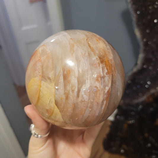 Hand holding a polished fire quartz sphere with orange and yellow inclusions against a blurred indoor background