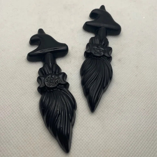 Two black obsidian broom-shaped figurines with witch hats and floral details on a white background