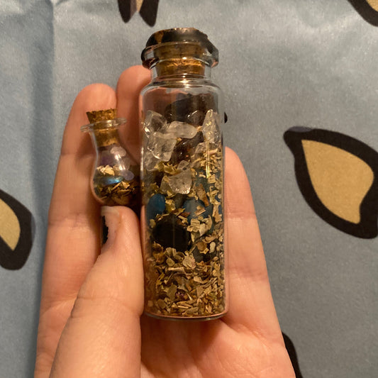 Hand holding two glass spell jars filled with herbs, crystals, and curiosities on a patterned fabric background